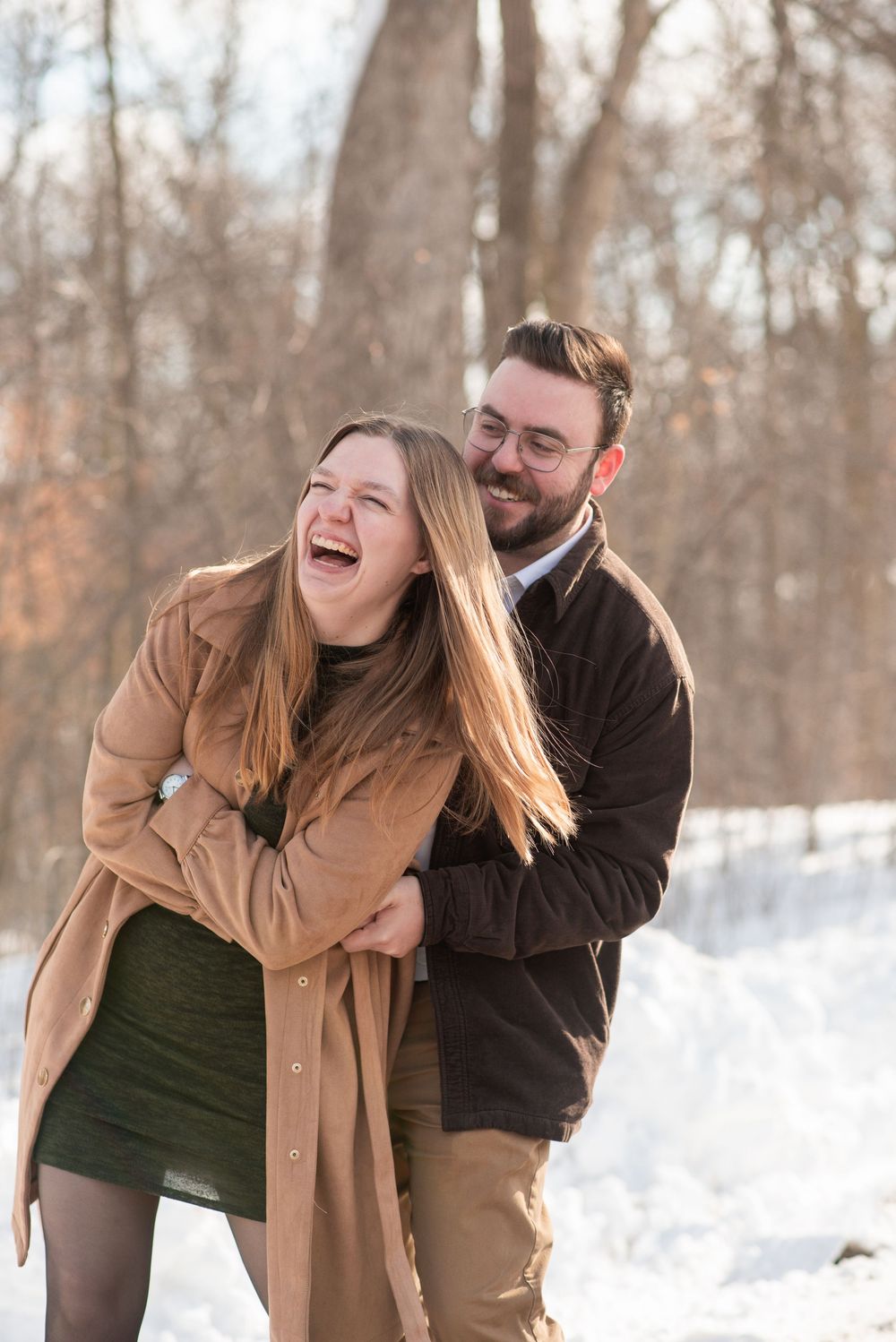 Couple laughing and embracing outdoors in winter snow, wearing warm coats among bare trees in golden sunlight.