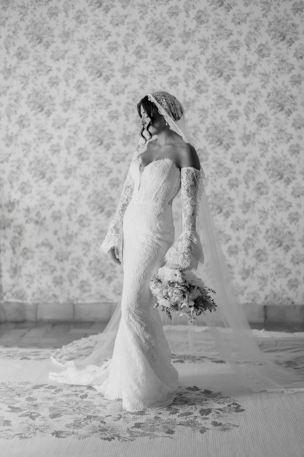 A bride in a classic lace wedding gown kneels gracefully in front of a floral wallpaper backdrop during a bridal session.