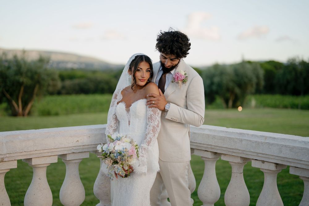 A romantic wedding couple embrace on an elegant stone balcony overlooking rolling green hills at sunset.