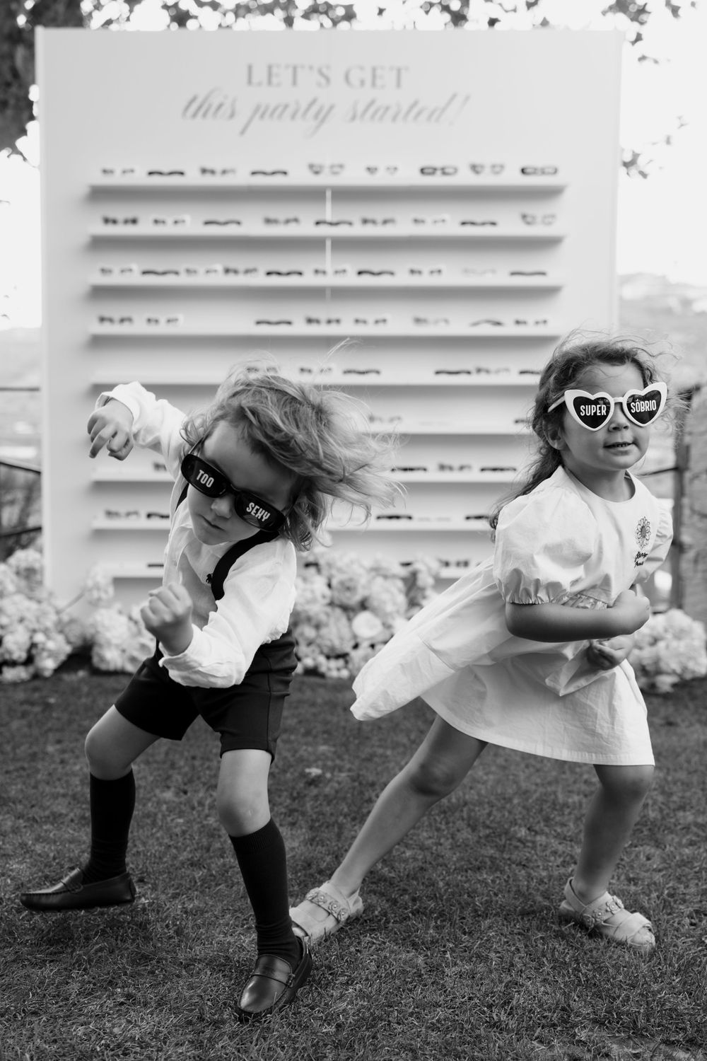 Two children in black and white leap across grass, wearing white garments and sunglasses in a playful fashion series.