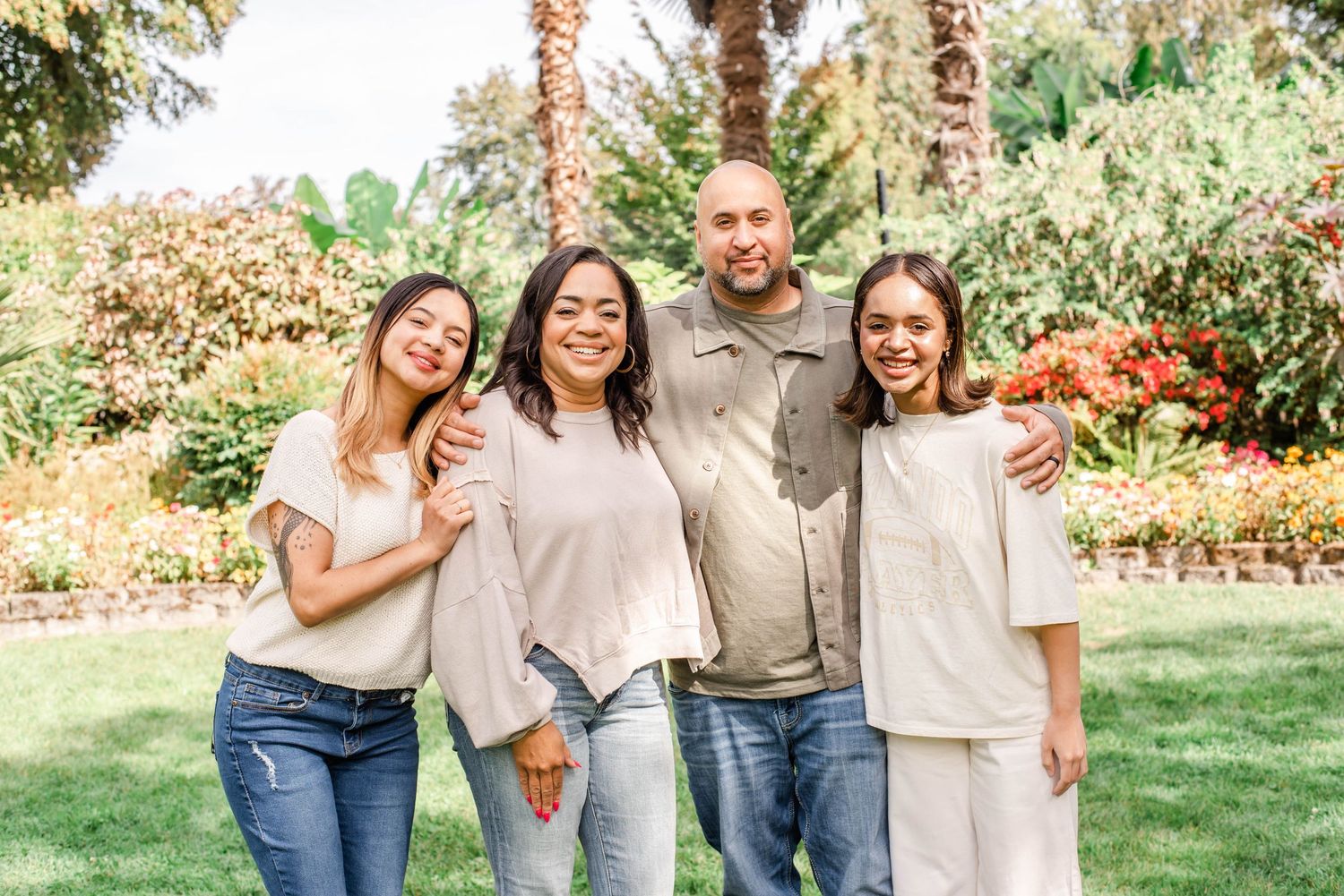 A group stands together smiling in a sunny garden with palm trees and colorful flowers in the background.