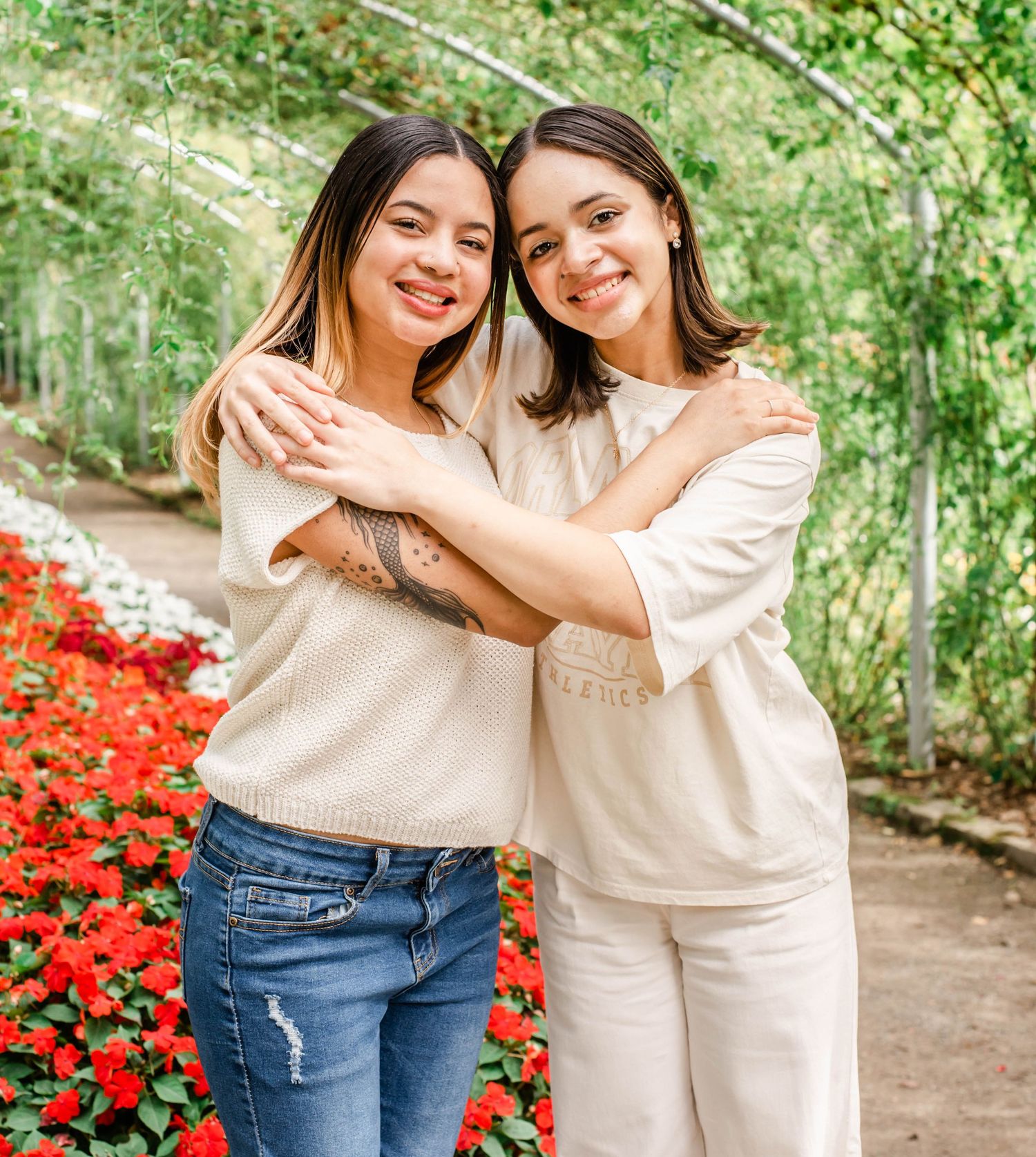 Two friends in light-colored tops embrace while standing near bright red flowers in a garden setting.