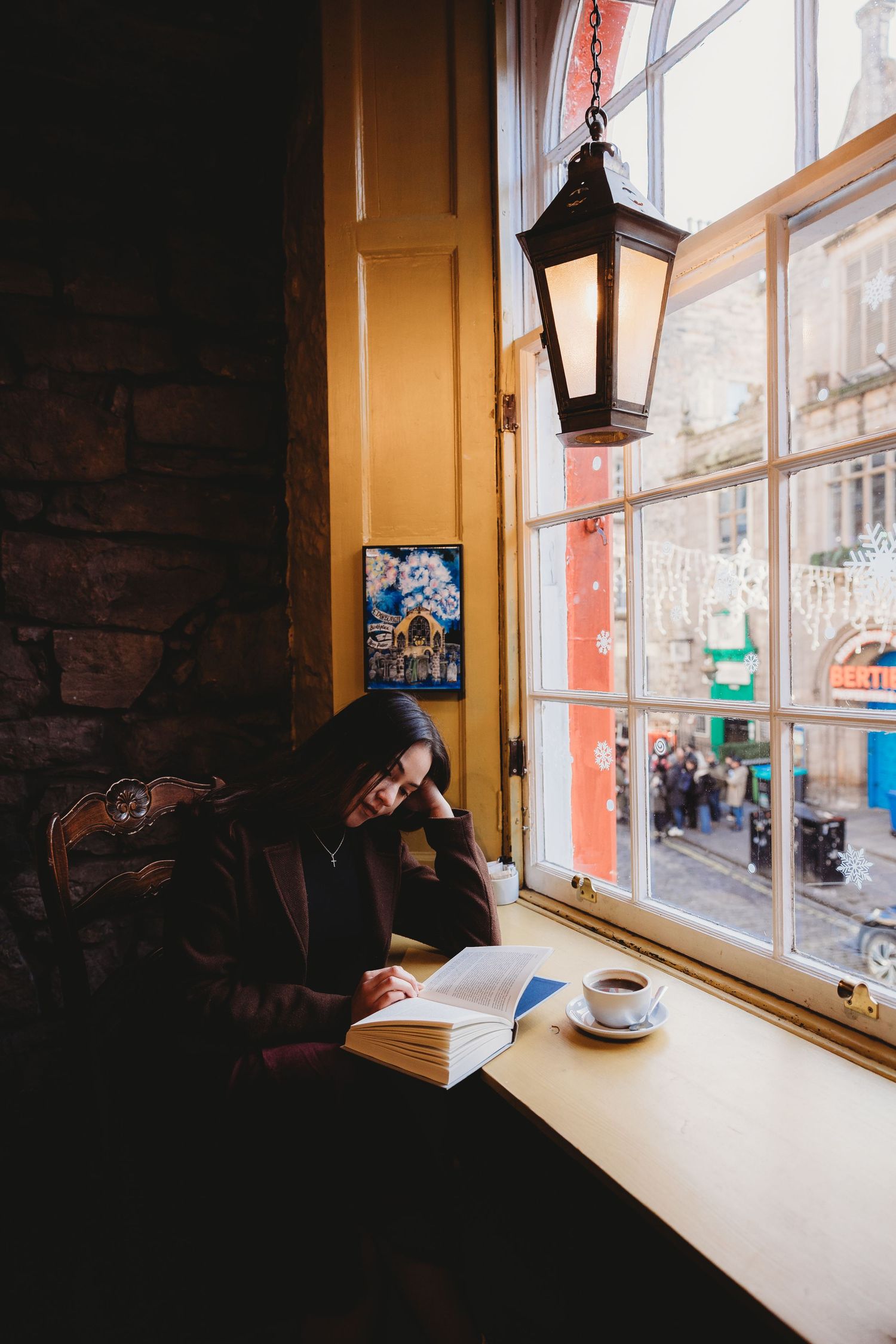 Cozy cafe window seat with coffee cup and book on wooden table overlooking city street through vintage windows.