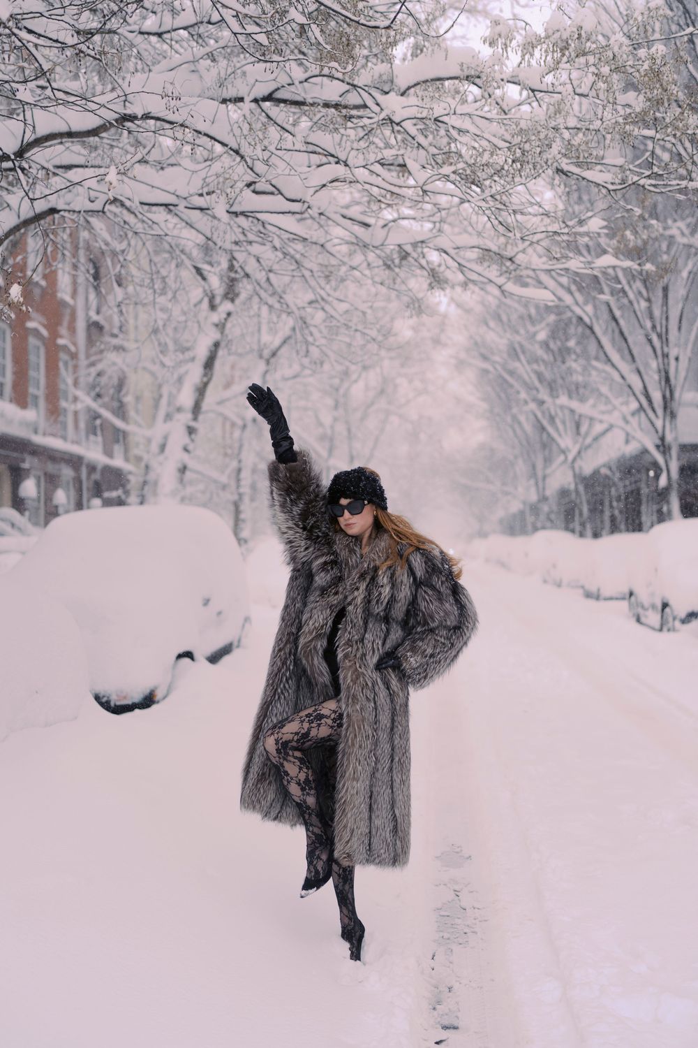 A figure in a luxurious fur coat and sunglasses dances joyfully in a snow-covered city street lined with trees.