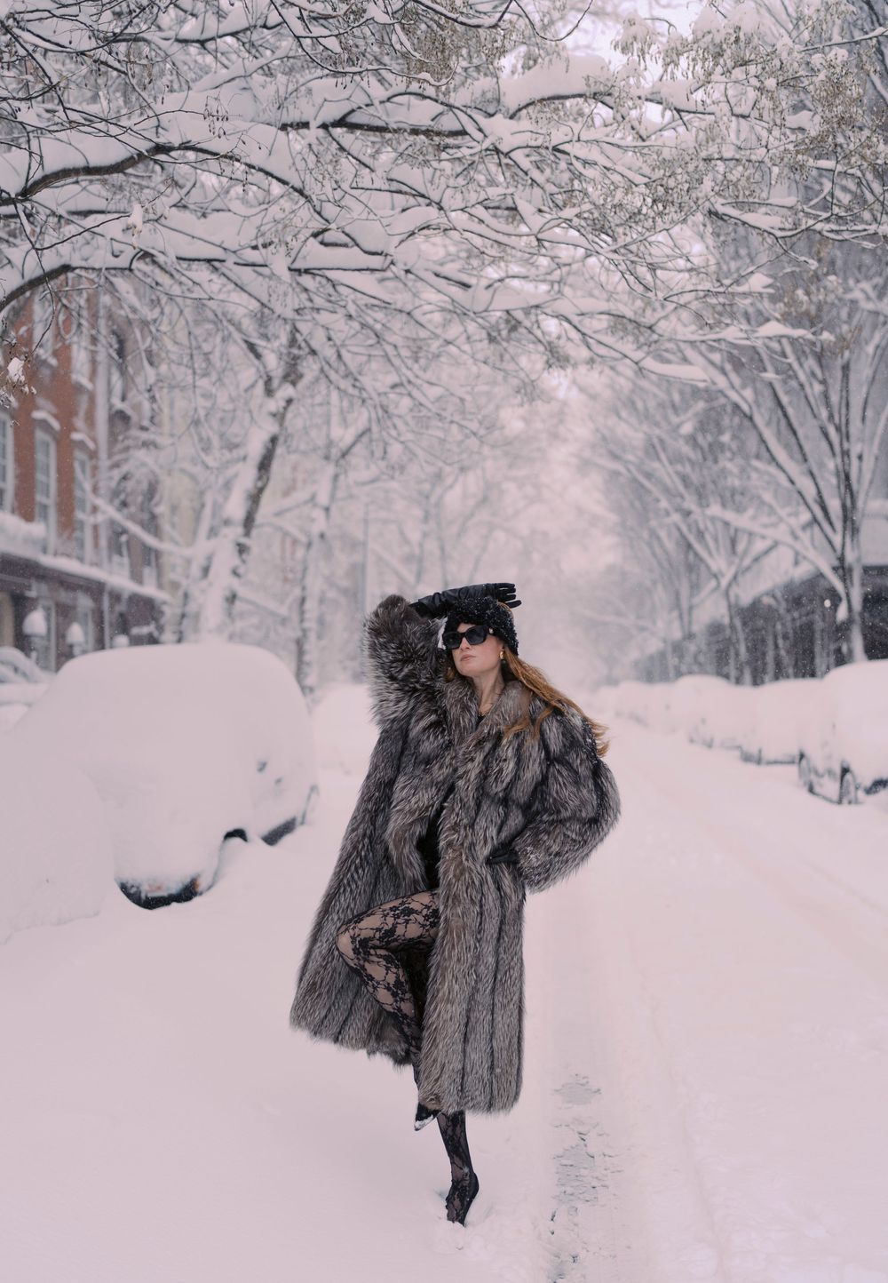 A person in a fur coat walks through a snowy winter wonderland with snow-covered trees lining the street.