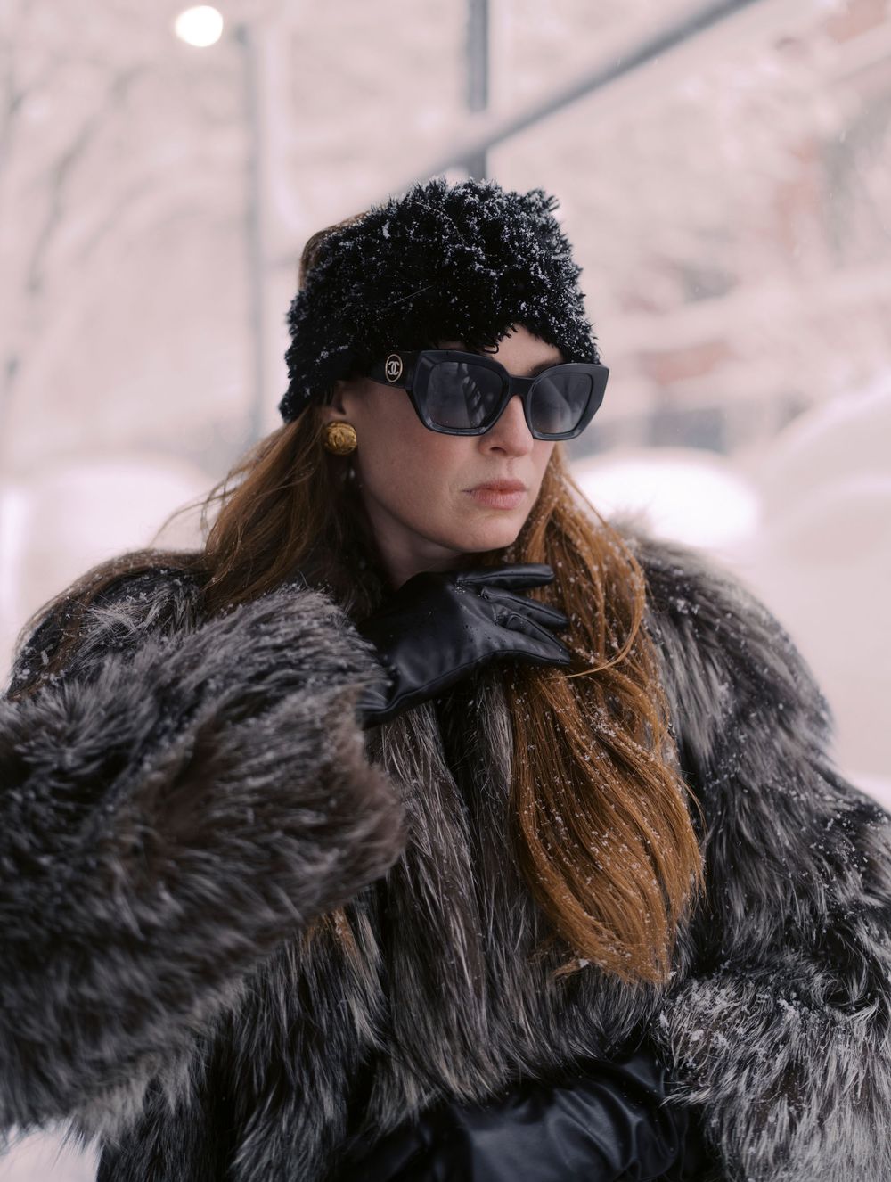 A person wearing sunglasses and a black fur coat poses in a winter setting with snowy background.