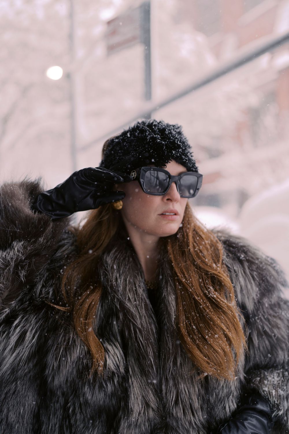 Person wearing sunglasses and a black fur coat poses in snowy winter weather with brown hair flowing in the wind.