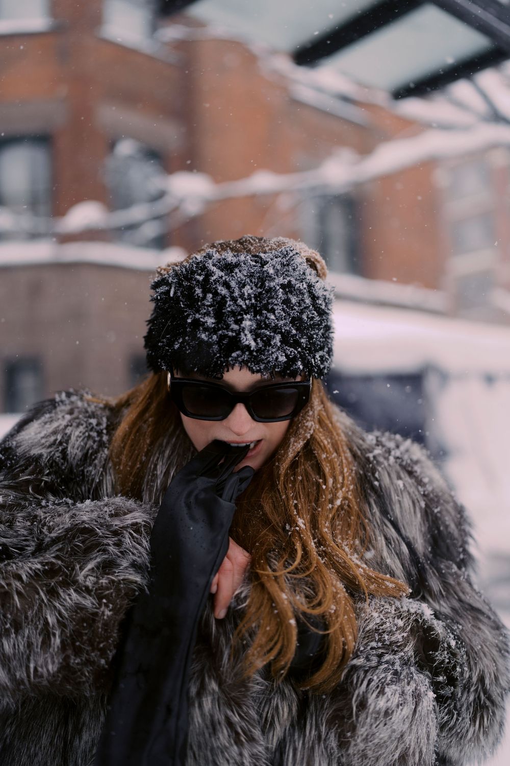 Person in dark winter coat and sunglasses with long brown hair stands against snowy urban backdrop.