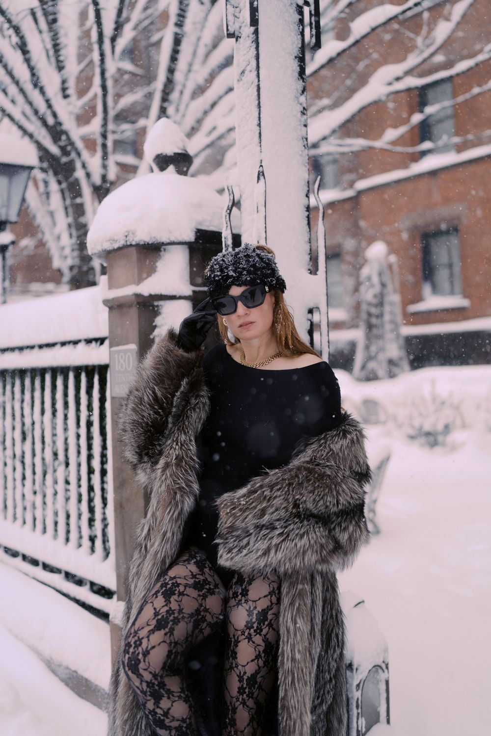 Person in a luxurious fur coat stands in a snowy winter setting with snow-covered trees and fences in the background.