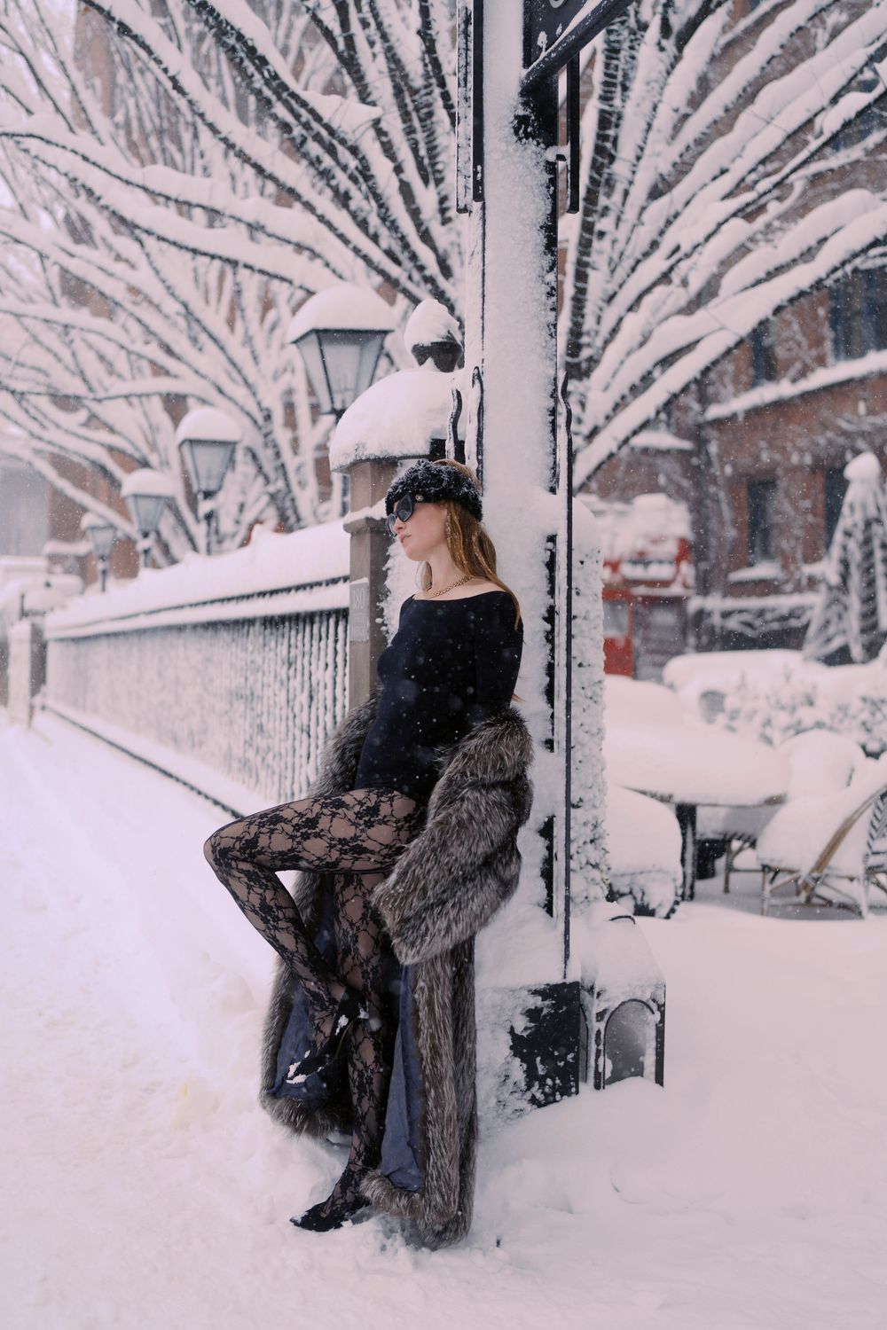 A figure in a dark coat sits on a snowy bench beneath snow-covered trees in a serene winter scene.