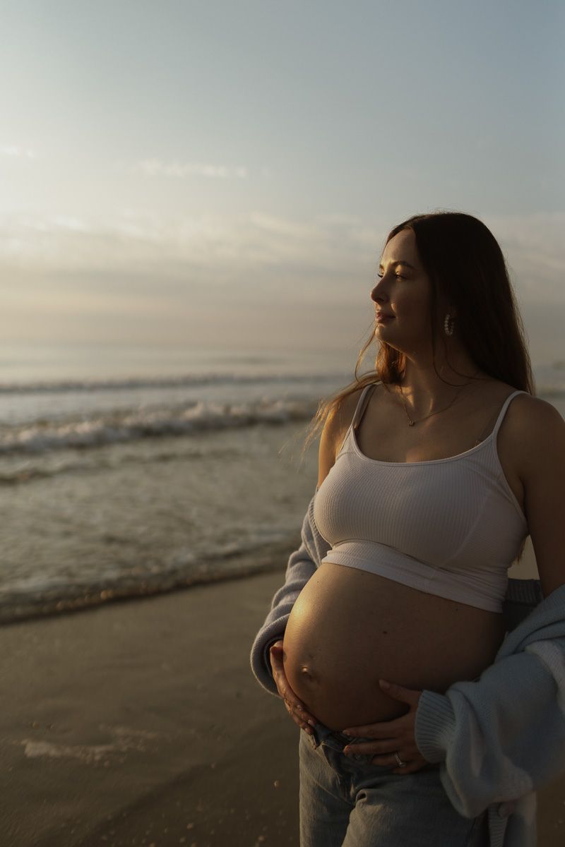 A pregnant woman in a white tank top sits peacefully on the beach at sunset, cradling her baby bump.
