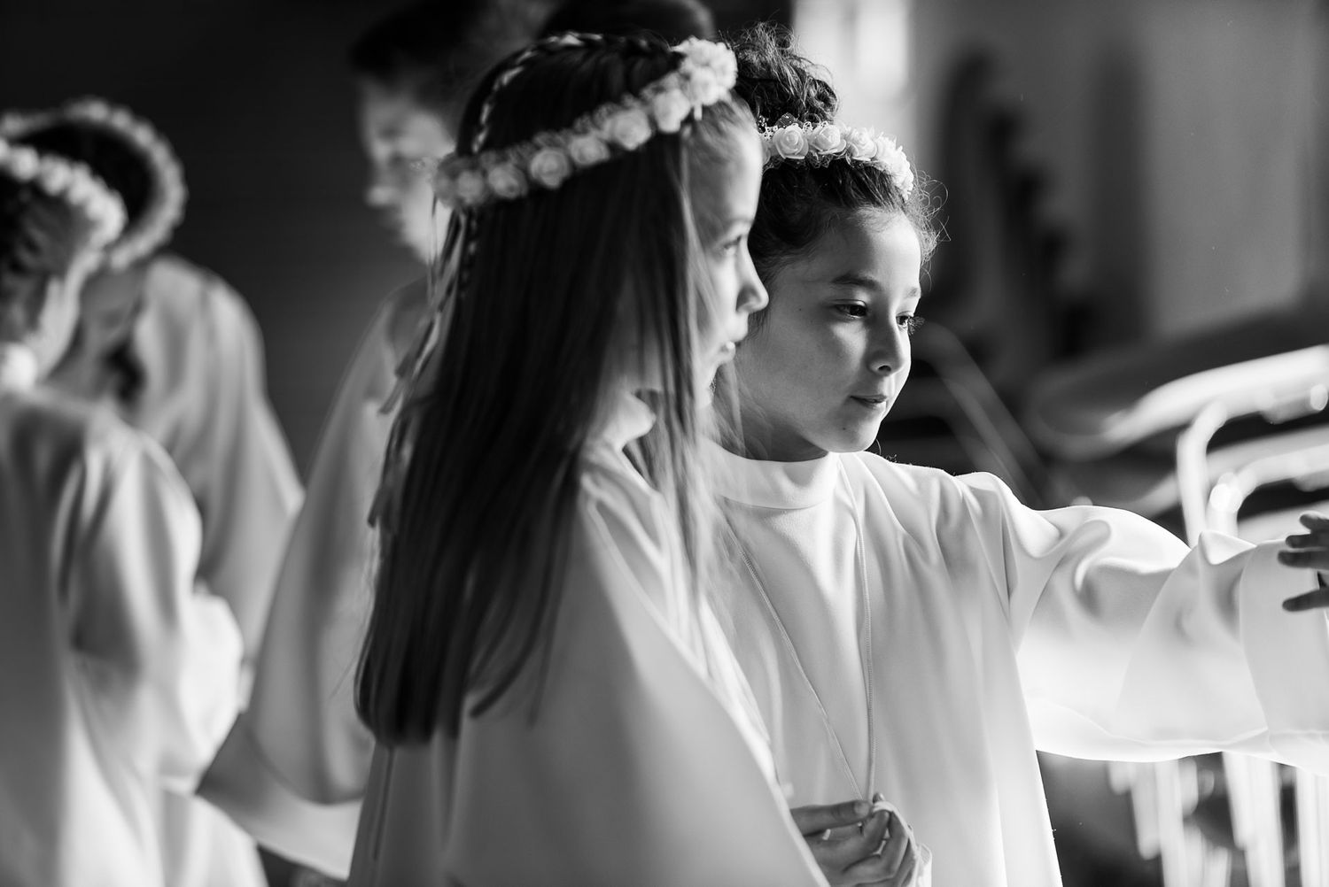 Children in white robes with floral crowns during religious ceremony.