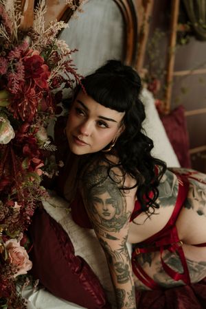 Vintage boudoir photo with burgundy floral arrangements, featuring dark curled hair and artistic tattoos in moody lighting.
