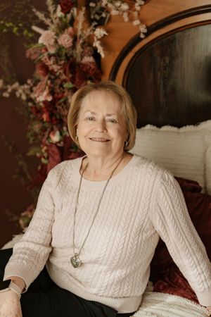 A person wearing a white cable knit sweater and heart necklace sits against a dark backdrop with floral decorations.