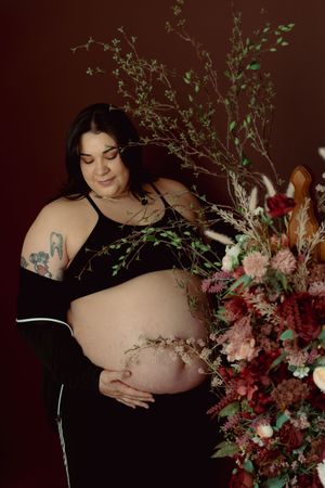 Artistic maternity photoshoot featuring dark backdrop with delicate pink and red floral arrangements.