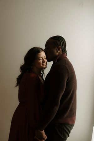 A romantic couple in brown sweaters embraces tenderly against a light neutral background in a series of intimate poses.