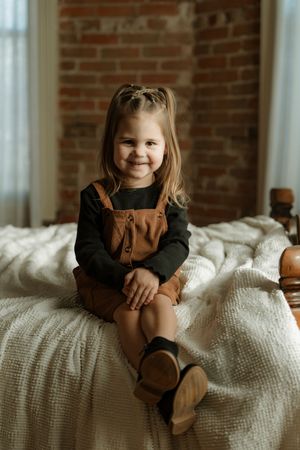 Child sitting on white knit blanket against exposed brick wall in brown and black clothing.