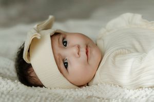 Baby wearing white headband lying on soft white knitted blanket in gentle lighting.