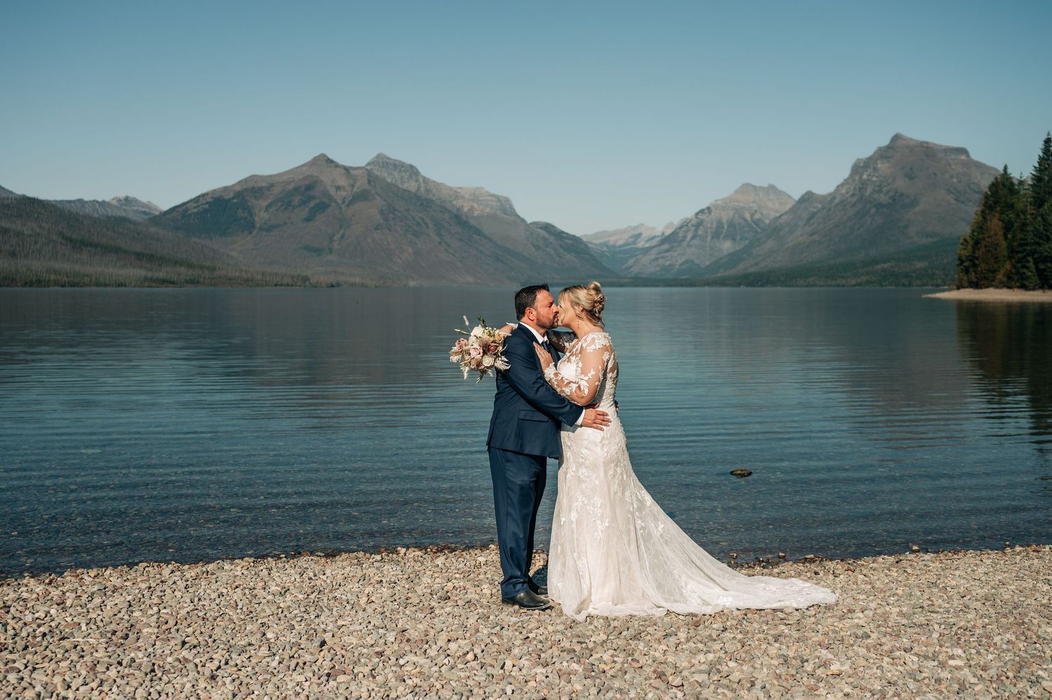 Scenic mountain lake wedding photos with couple embracing on rocky beach at sunset with dramatic peaks in background.
