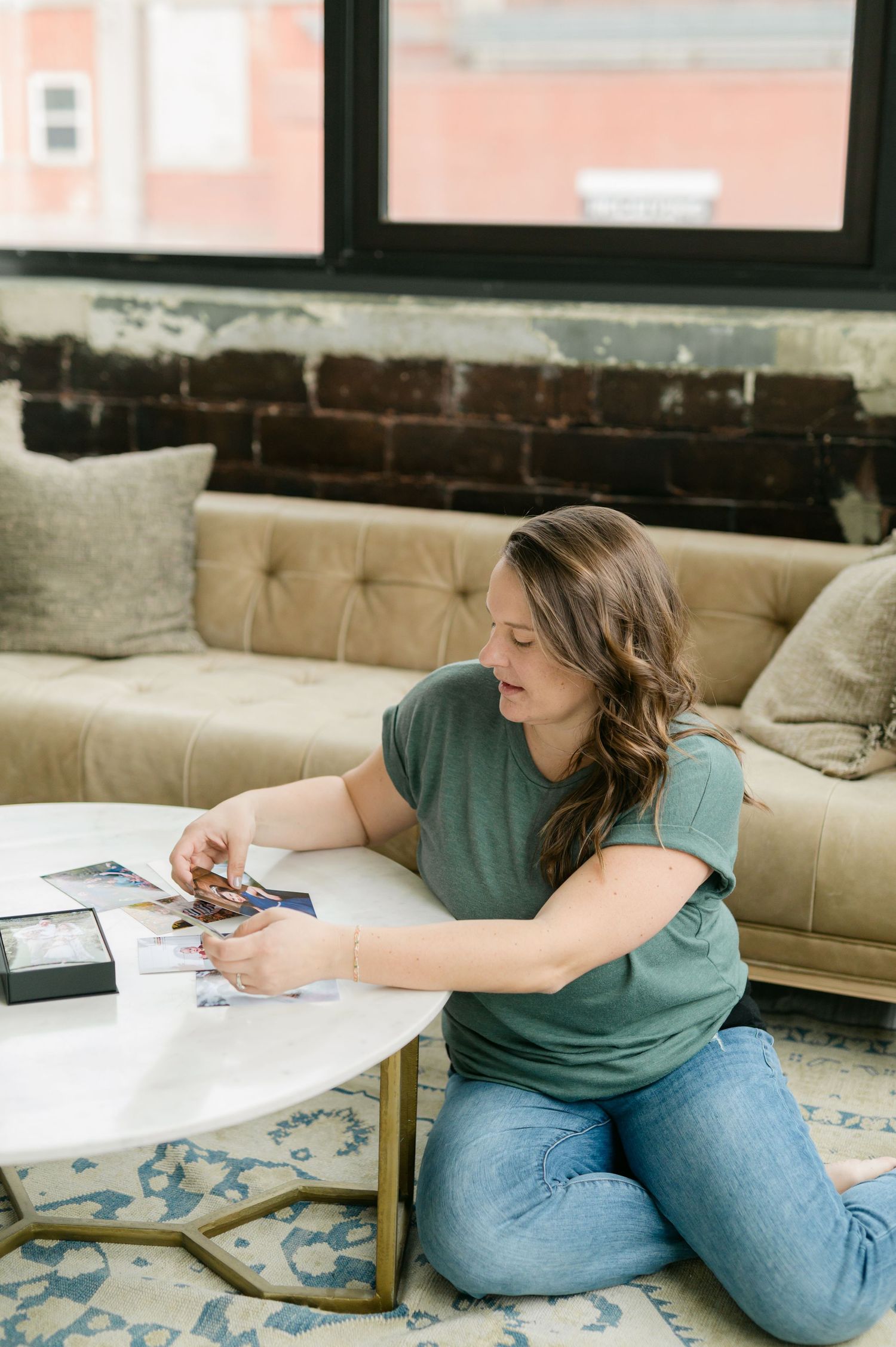 Artist in teal shirt and jeans sketches while sitting on beige couch with patterned blue rug below.