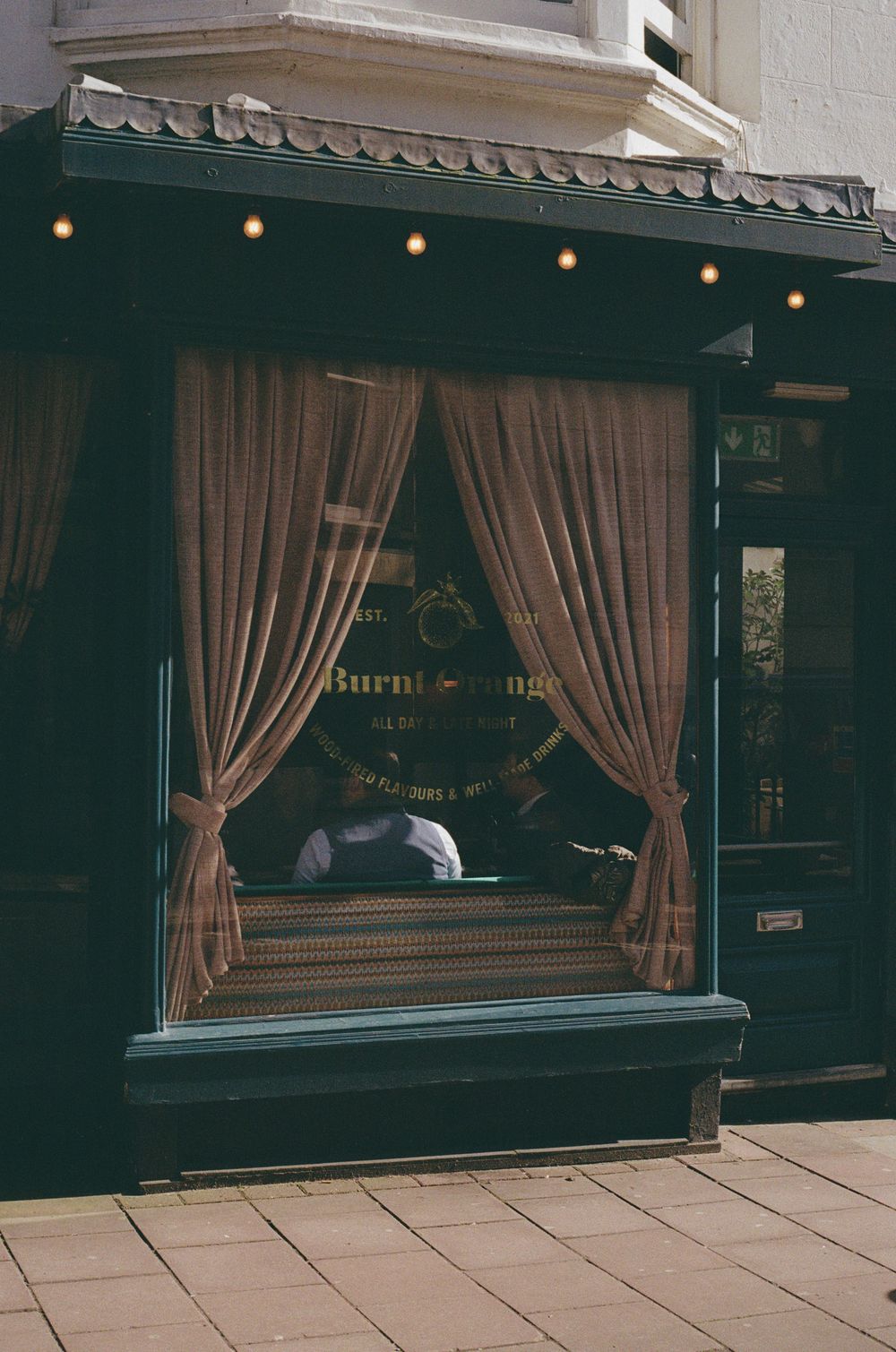 A row of dark green storefronts with beige curtains and vintage-style windows along a stone paved street.