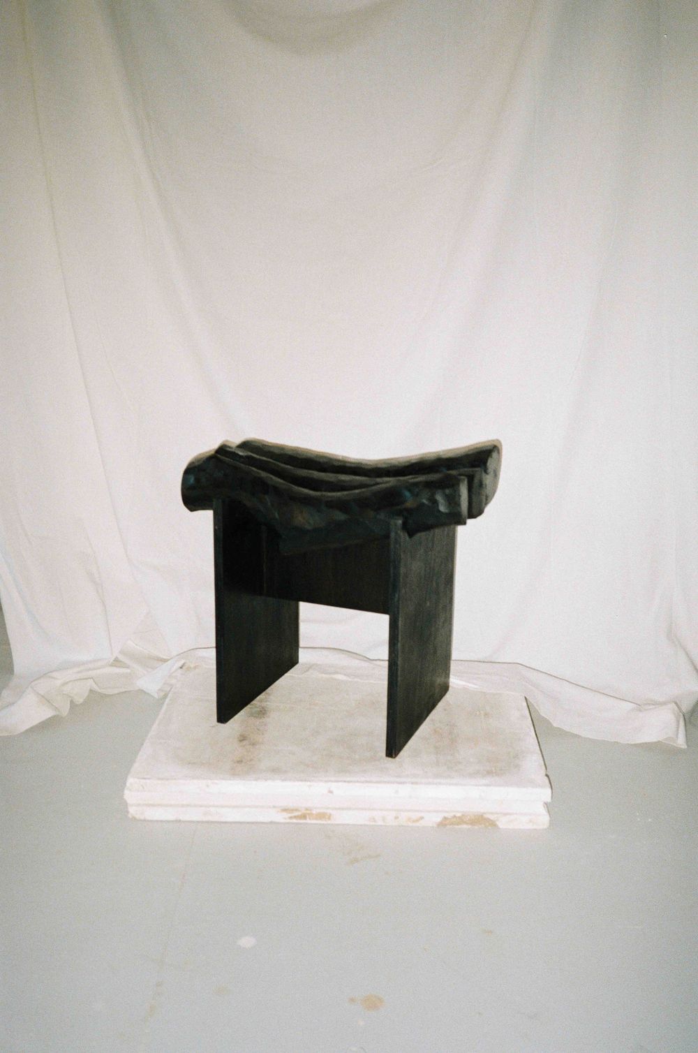 Dark sculptural wooden stool with draped fabric seat displayed on white platform against white backdrop.
