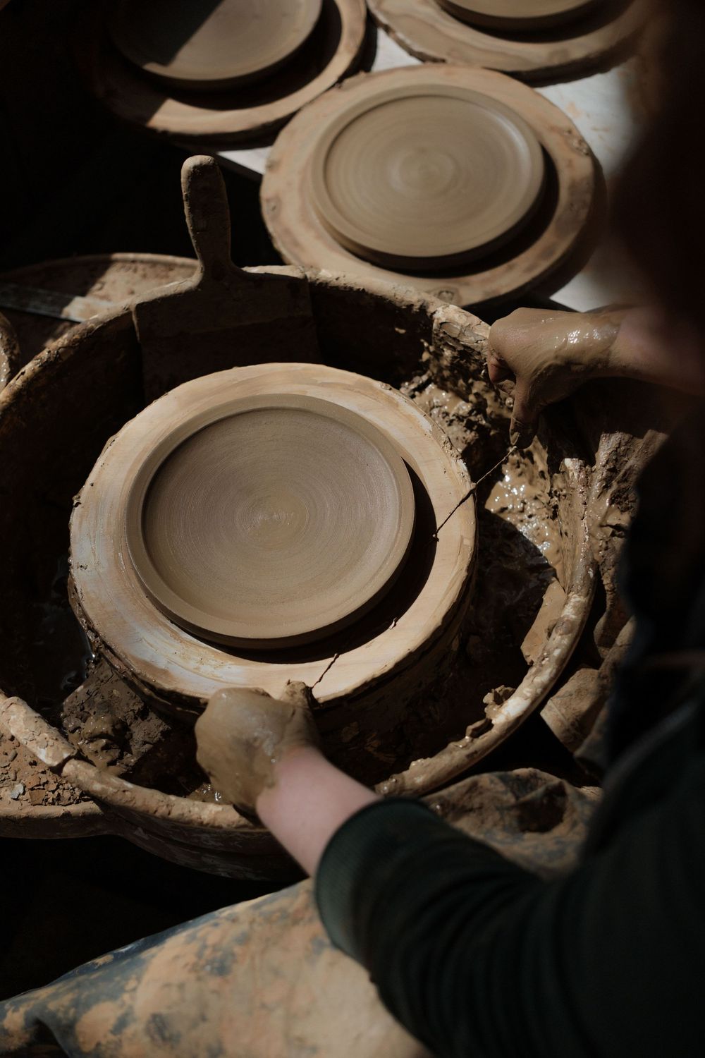 Potter shaping clay on a spinning wheel, surrounded by freshly made ceramic plates in a rustic workshop.