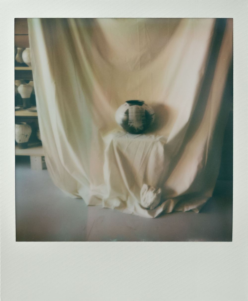 A dark ceramic vase displayed against a draped white fabric backdrop with pottery shelves visible in the background.