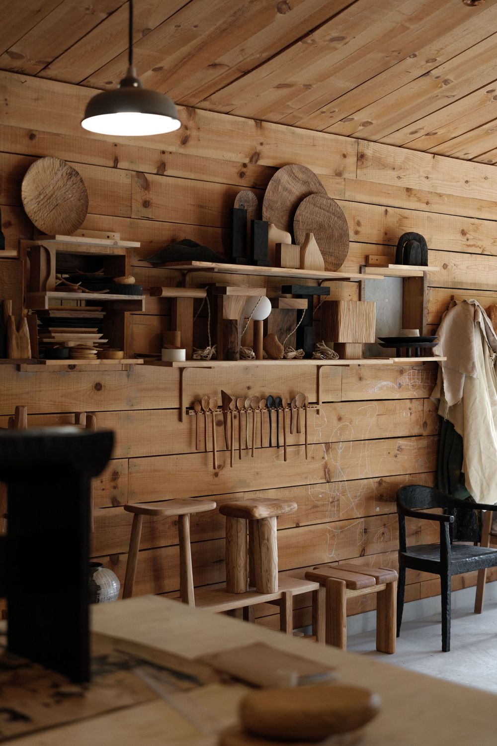 Rustic wooden carpentry workshop with handmade stools, wall-mounted tools, carved wooden pieces on shelves, and a pendant light.