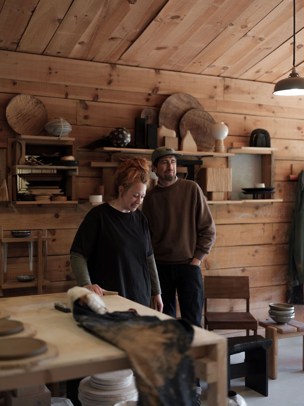 Two artisans work in a rustic wood-paneled pottery studio with shelves displaying ceramic pieces and handcrafted goods.