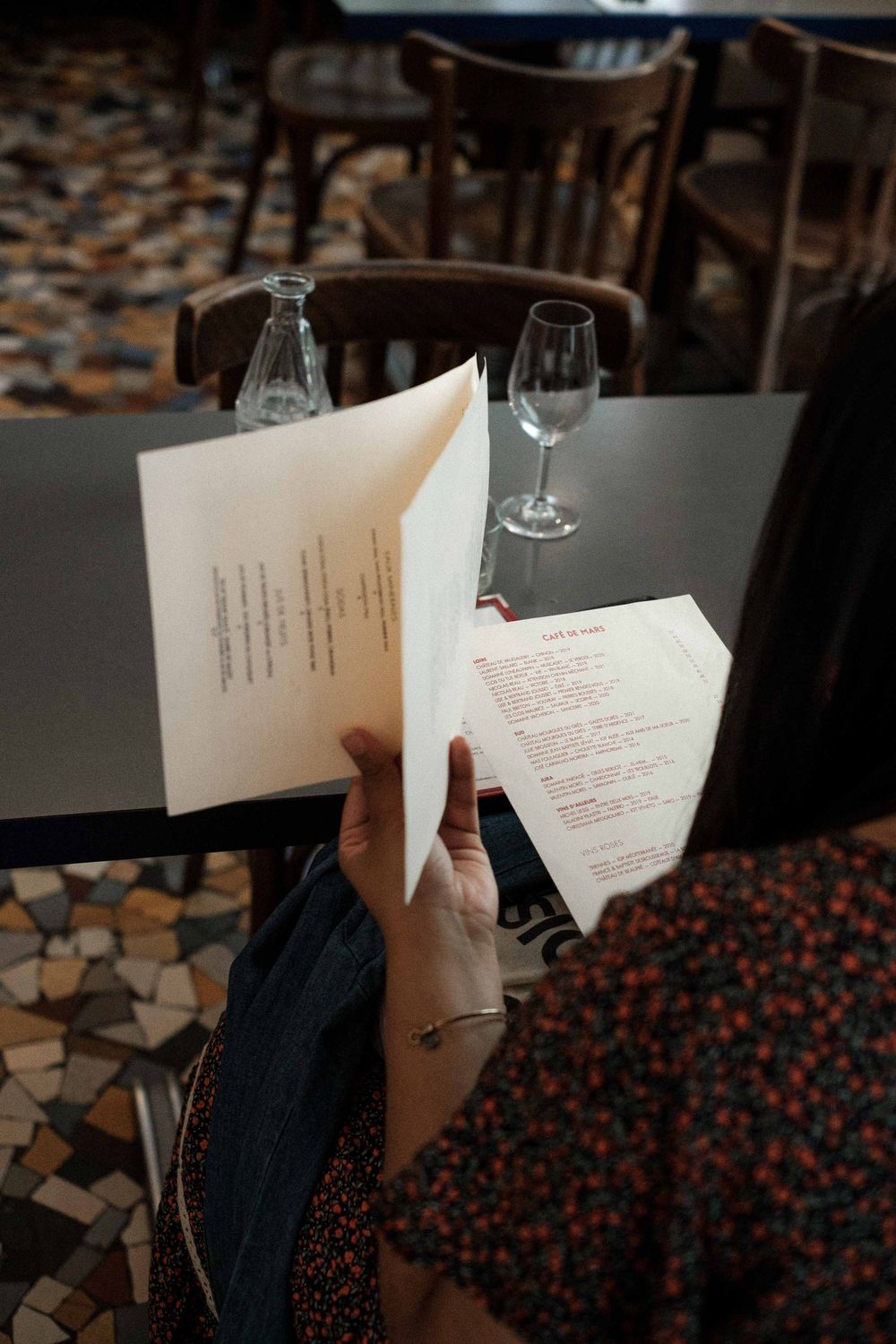 A restaurant menu being held over a table with a wine glass and patterned floor visible in the background.
