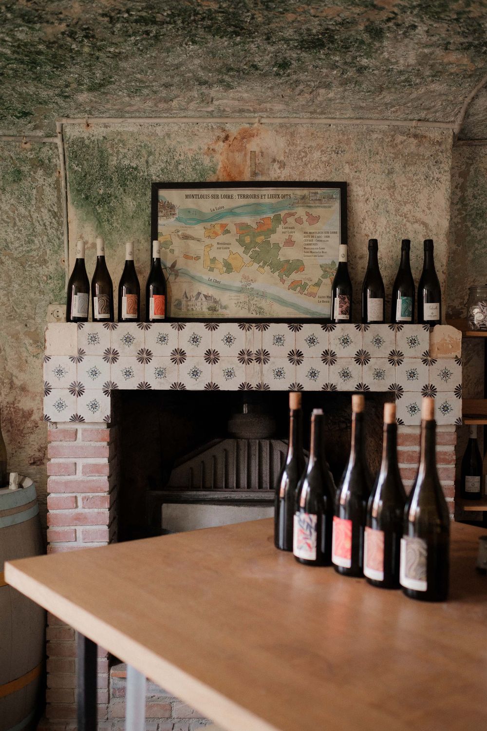 Wine bottles displayed on a rustic brick mantel with a framed map above and more bottles on a wooden table below.