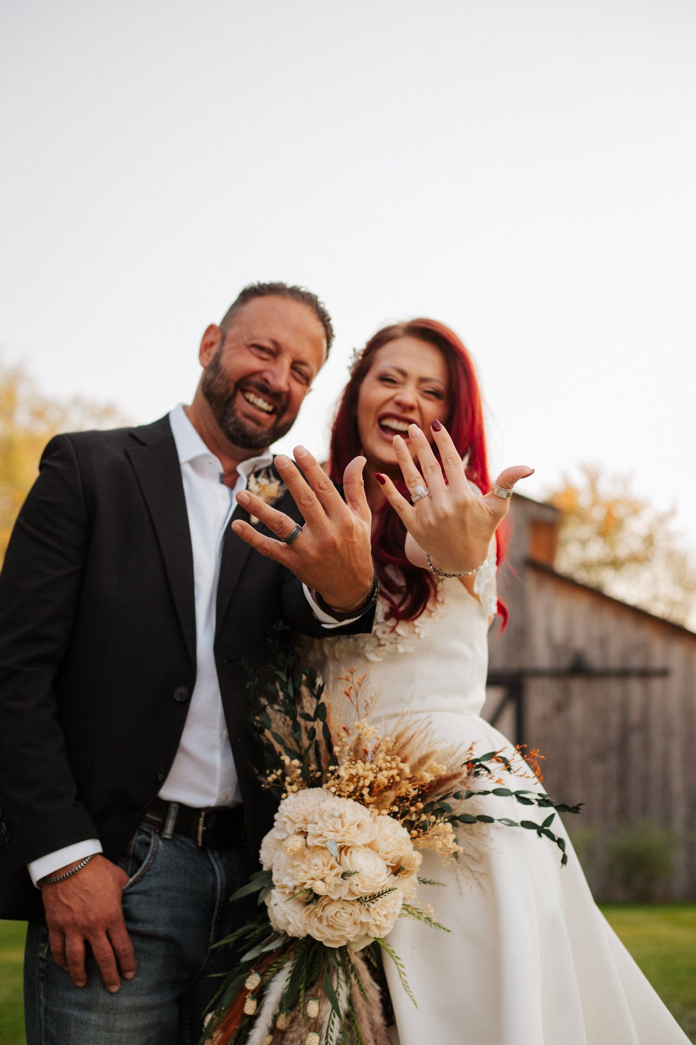 Joyful newlyweds showing off wedding rings with bouquet of white flowers outdoors.