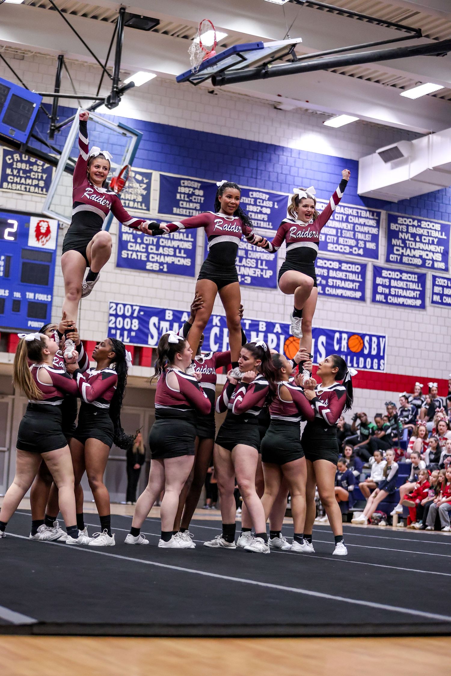 Cheerleaders in navy blue uniforms perform synchronized stunts during a routine in a school gymnasium.