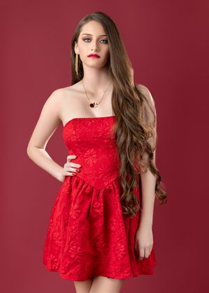Woman in strapless red lace dress poses against burgundy backdrop in fashion photography series.