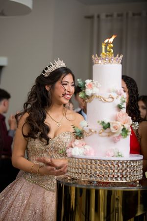 Elegant tiered pink and white quinceañera cake displayed on gold stand with floral decorations and number 15 candle on top.