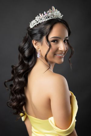A person wearing a yellow strapless dress and sparkling tiara poses elegantly against a dark background.