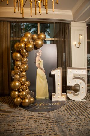 Elegant party setup with illuminated number 15 display and gold balloon arch against dark wall with textured carpet.