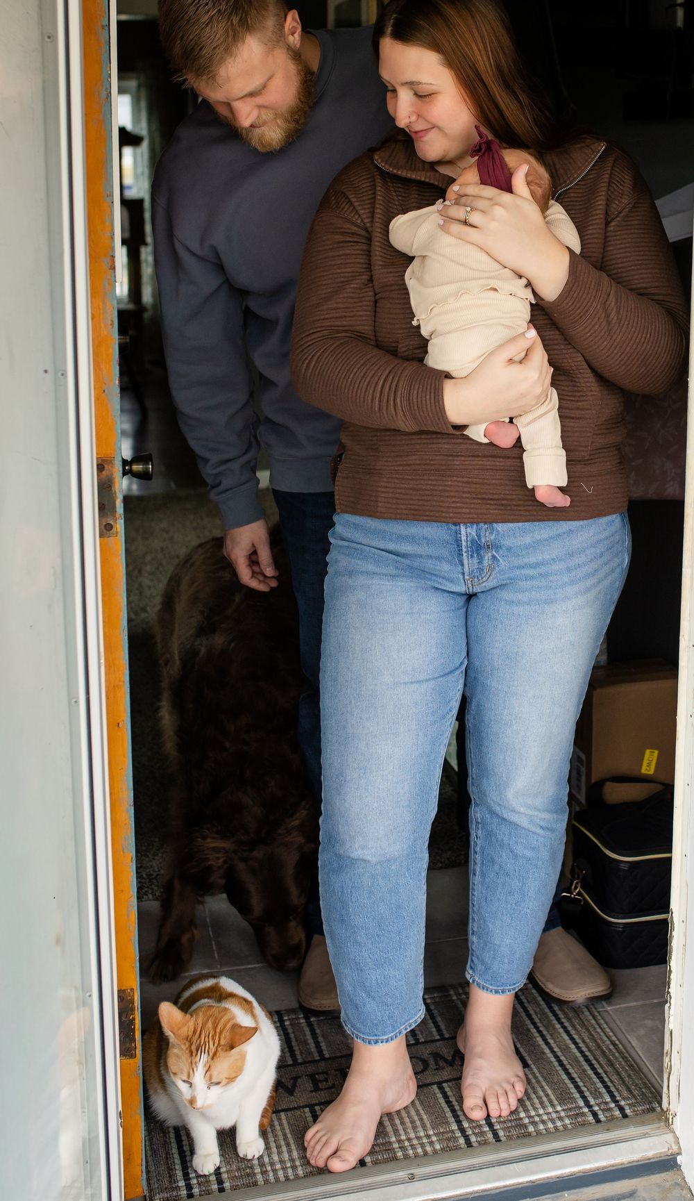 A cat sits in a doorway watching someone holding a baby while wearing blue jeans and a brown sweater.