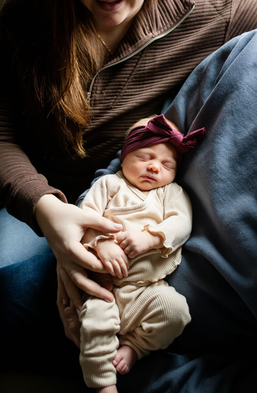 Sleeping newborn baby wearing cream colored outfit and burgundy bow headband rests peacefully in caregiver's arms.