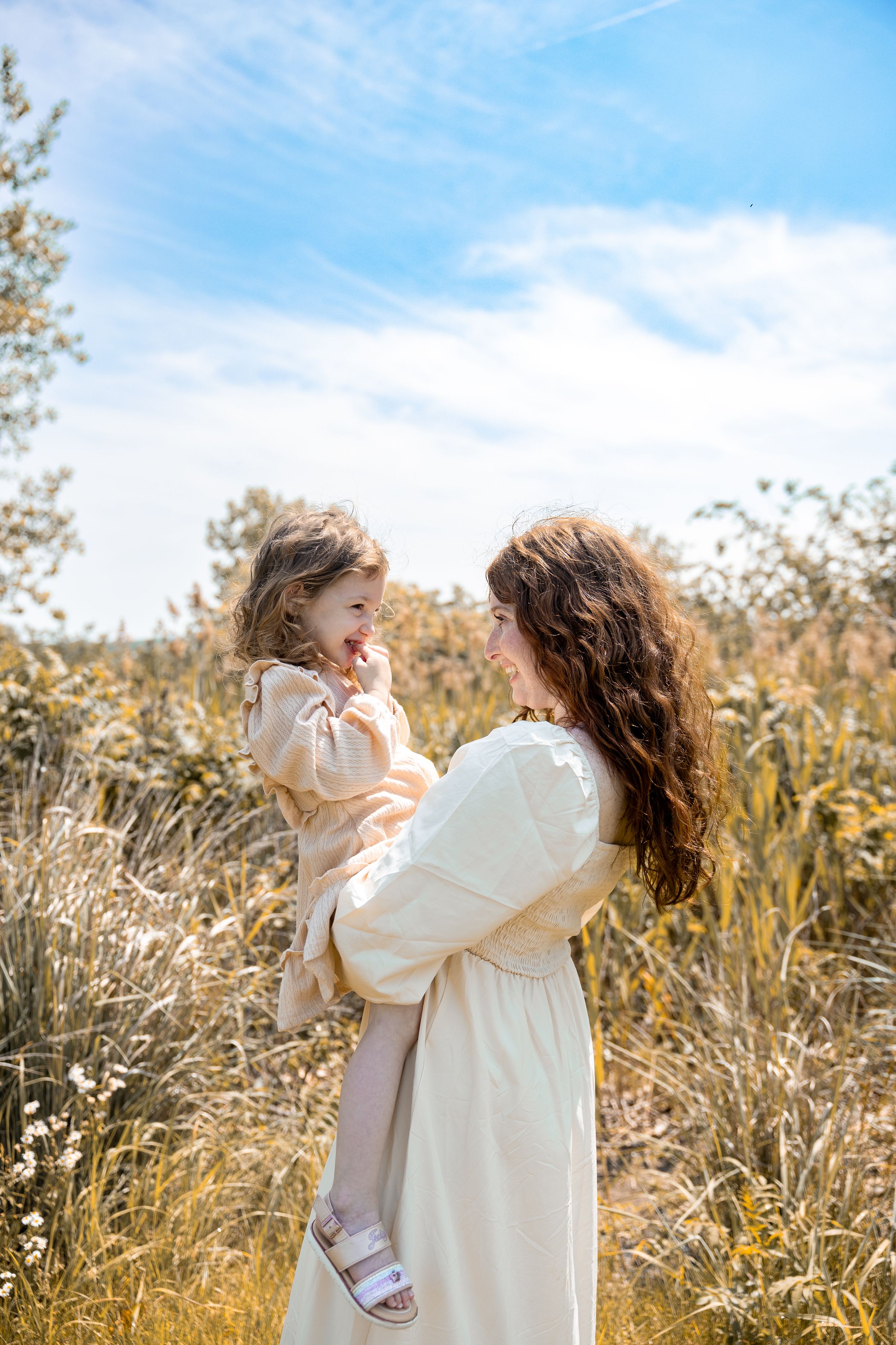 Two people embrace joyfully in a sunlit meadow with tall golden grass and blue sky in the background.