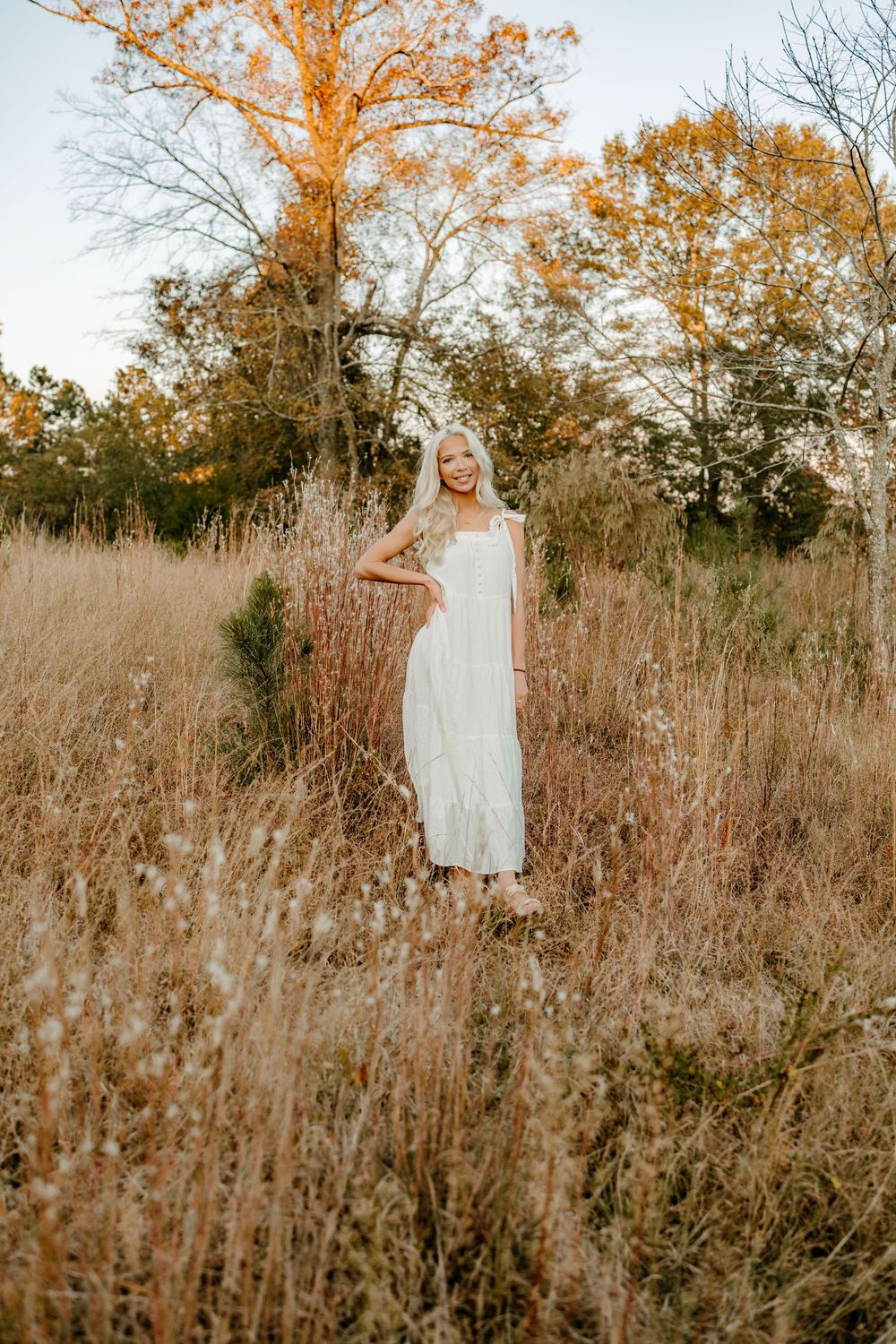 A white dress flows in the golden autumn field with tall dried grass and trees at sunset.