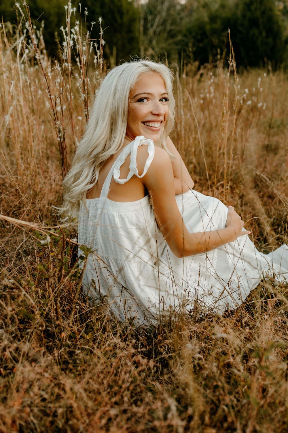A bride in a white lace dress poses gracefully in a golden autumn field during sunset.