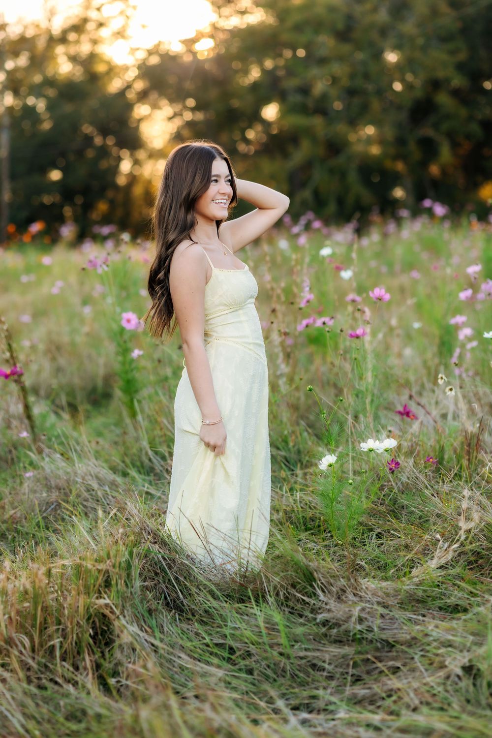Woman in pale yellow dress stands among tall grass and pink wildflowers at sunset.
