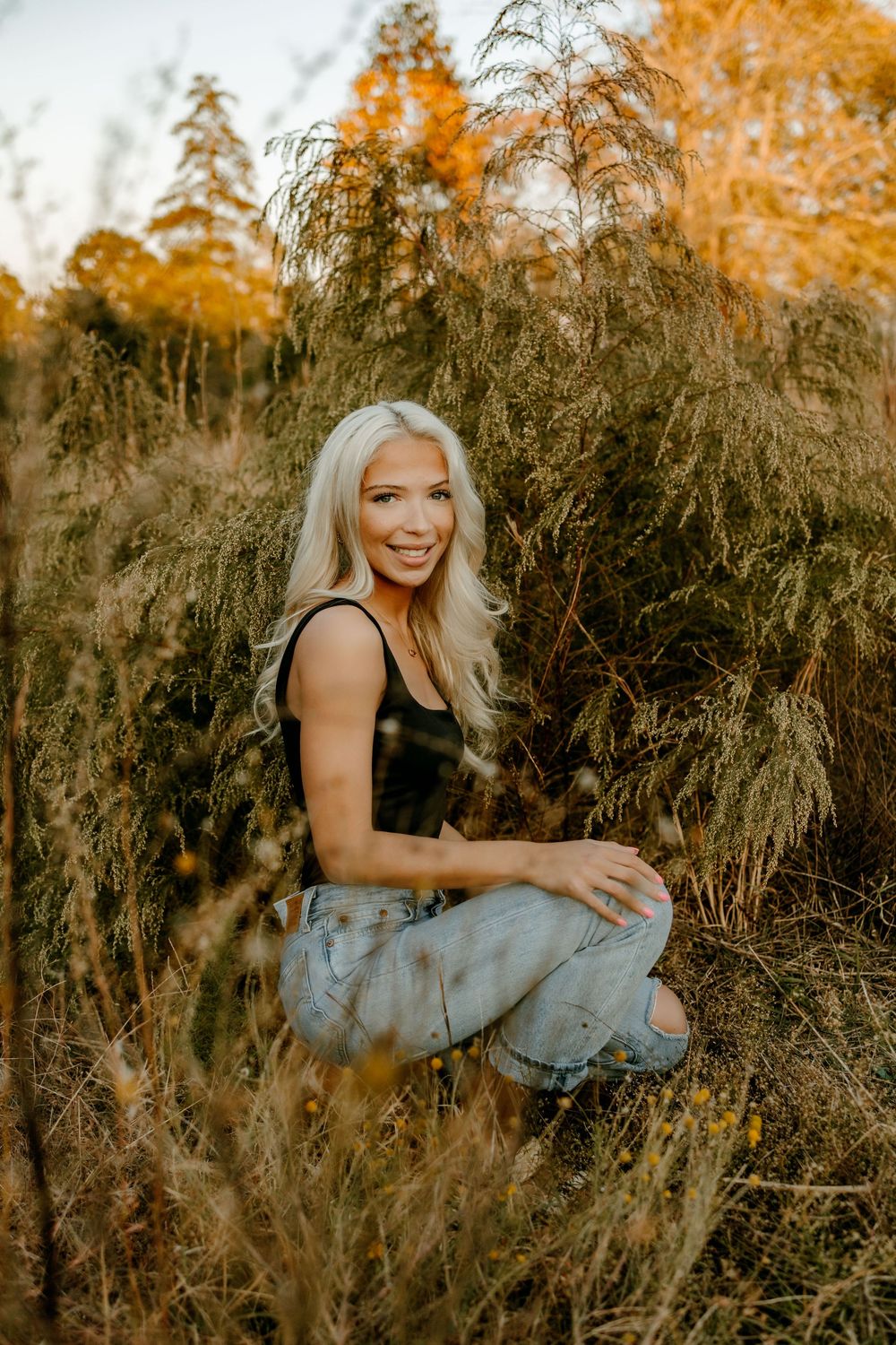 A casual autumn portrait among tall dried grasses with warm orange foliage in background.