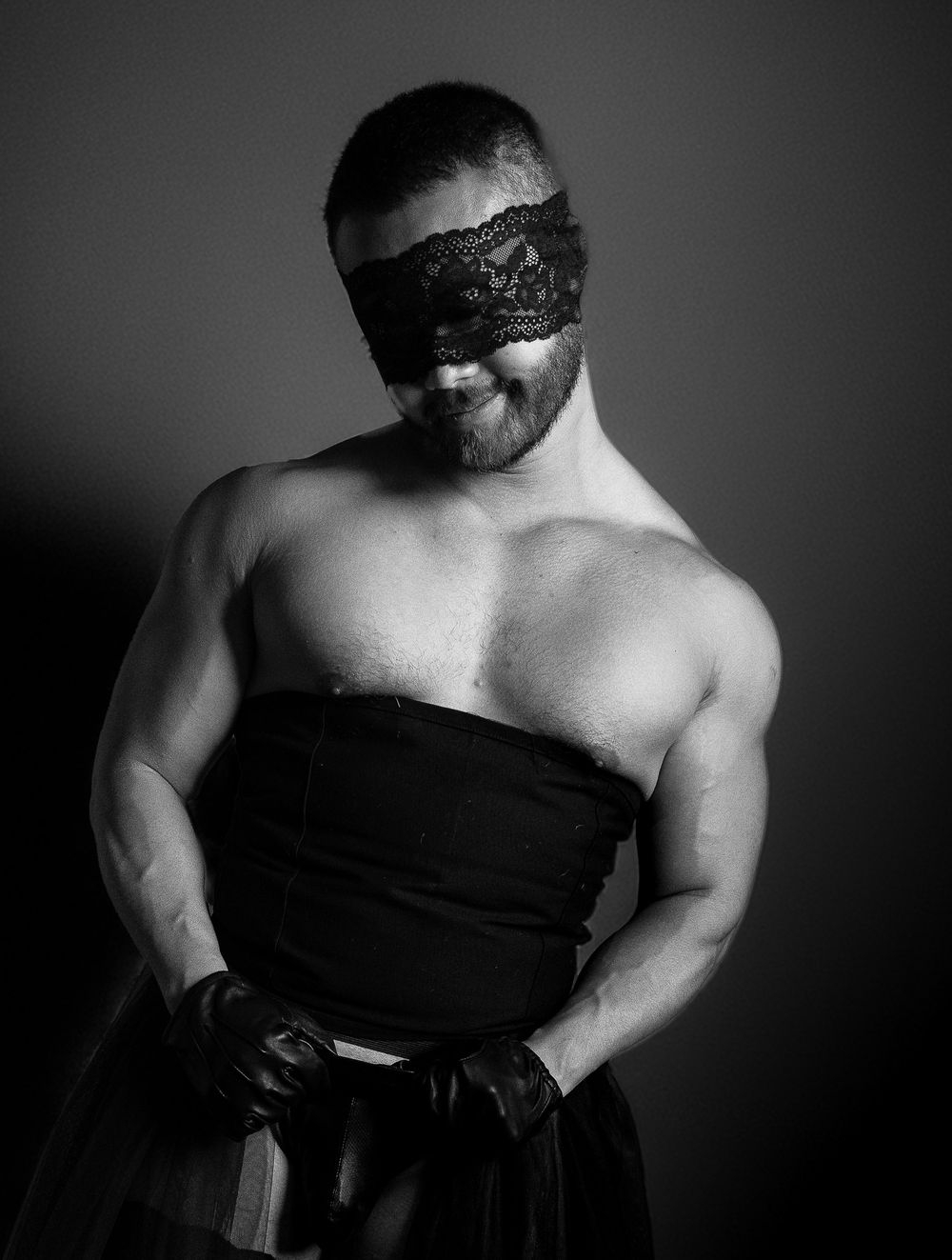 Black and white photo of a muscular bearded man wearing a lace blindfold and black strapless outfit.