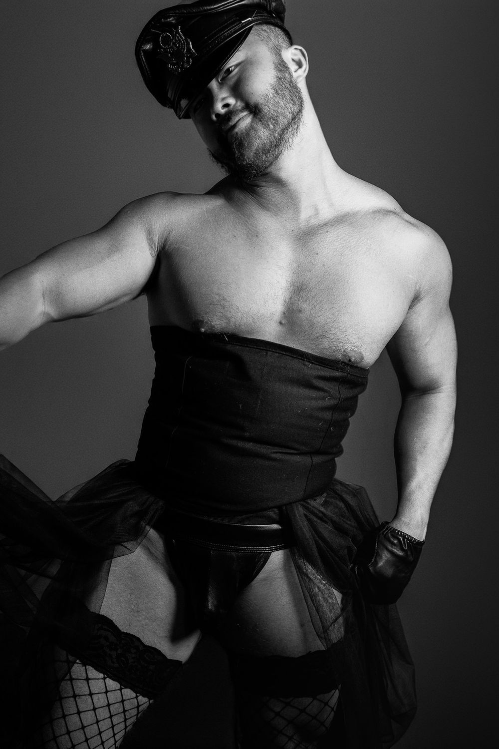 Black and white photo of a muscular bearded man in a leather cap, corset, and fishnet stockings.