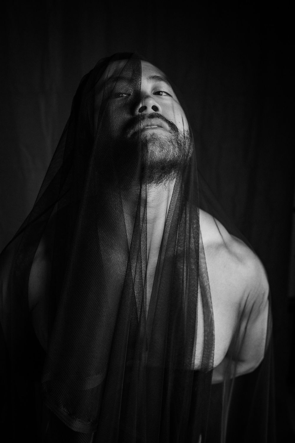 Dramatic black and white portrait of a bearded man with sheer black veil draped over his face.
