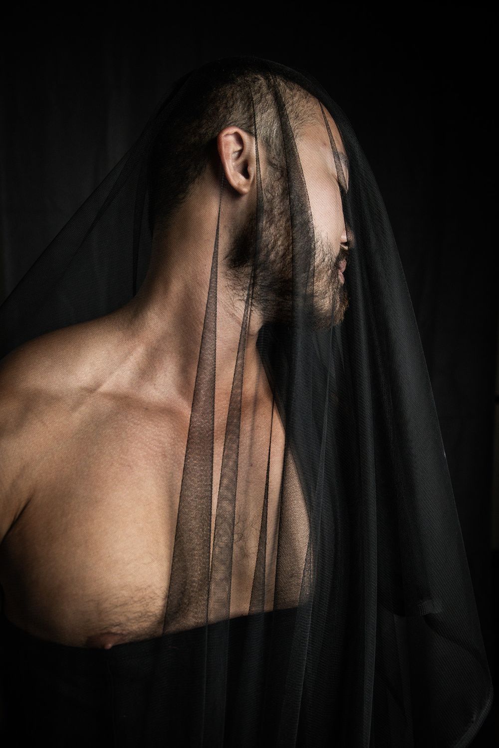 Shirtless bearded man draped in black sheer veil in dramatic dark artistic portrait.