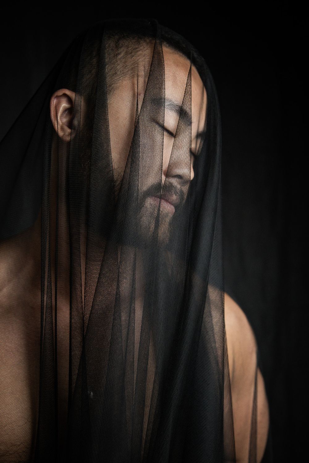 Moody dark portrait of a bearded man with sheer black fabric draped across his face, eyes closed.